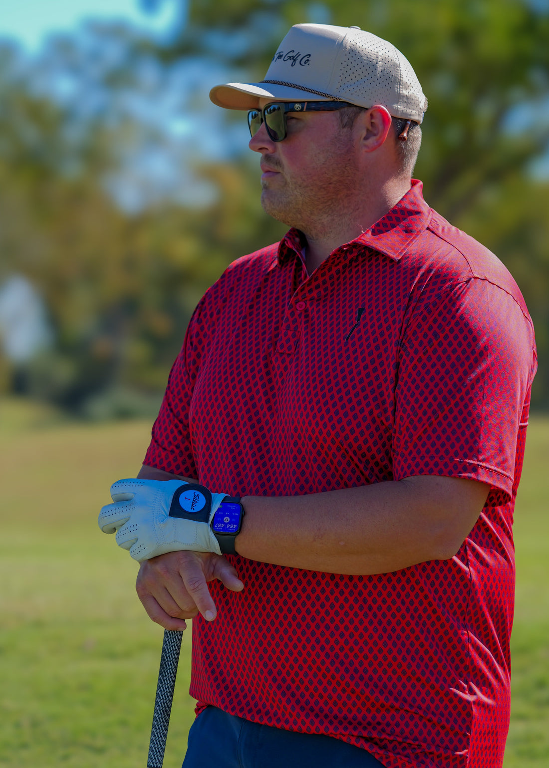 Man in a red shirt and tan cap holding a golf club on a golf course